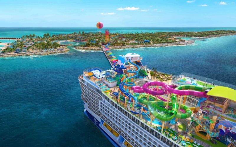 A vibrant aerial view of a Royal Caribbean cruise ship, featuring an elaborate top deck with colorful water slides and leisure areas, approaching the tropical island Coco Cay with clear blue waters and sandy beaches, under a bright blue sky.