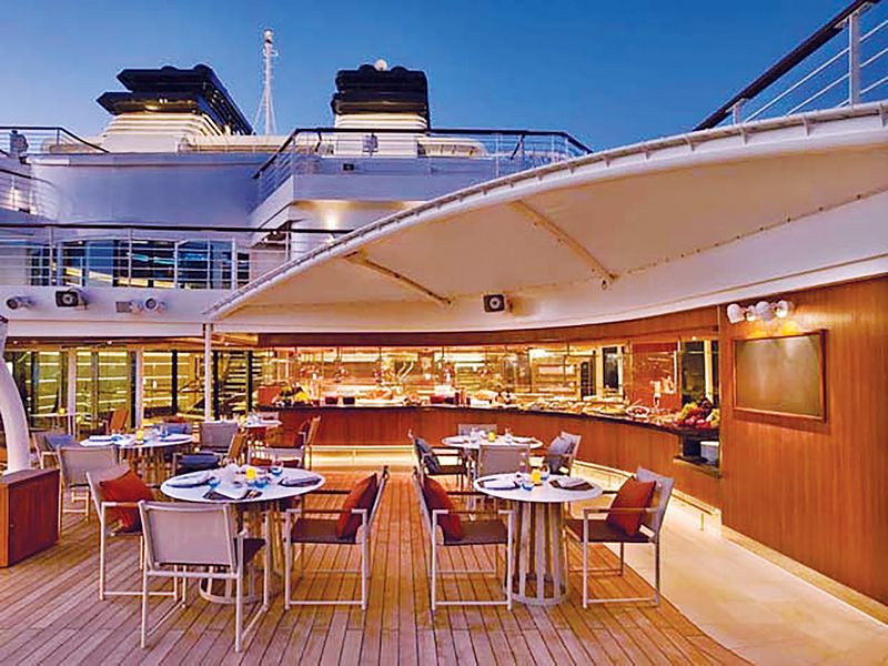 The Patio on a Seabourn cruise ship offers an al fresco dining experience with casual outdoor seating, set against the backdrop of a warmly lit buffet area and the open evening sky, reflecting a relaxed and inviting maritime atmosphere.