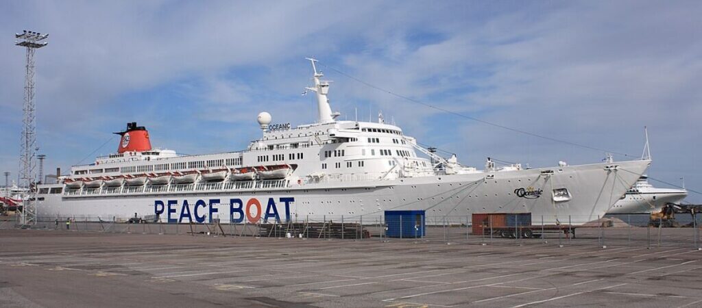 The SS Oceanic cruise ship docked in Helsinki, under a clear blue sky with few clouds. The ship is painted white with a distinctive red funnel and prominently displays the "Peace Boat" logo along its side. The photograph captures the ship in a spacious port area, surrounded by a large, empty parking lot and industrial equipment, highlighting the ship's readiness for embarking on its next voyage. The setting suggests a moment of calm before the activity typically associated with cruise departures.