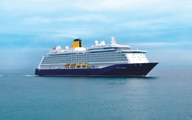 Spirit of Adventure gracefully sailing across calm blue waters. The vessel is modern and sleek, painted in a dark navy blue with a contrasting yellow funnel. Multiple decks lined with balconies are visible, and the ship is equipped with several large satellite domes. The overcast sky adds a serene and tranquil ambiance to the scene, emphasizing the ship's smooth voyage.