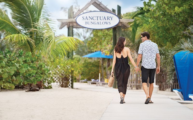 A couple walks hand in hand towards the Sanctuary Bungalows at Princess Cays, surrounded by lush tropical foliage. The relaxed ambiance is evident in their casual beachwear and the serene setting, promising a private and tranquil retreat.