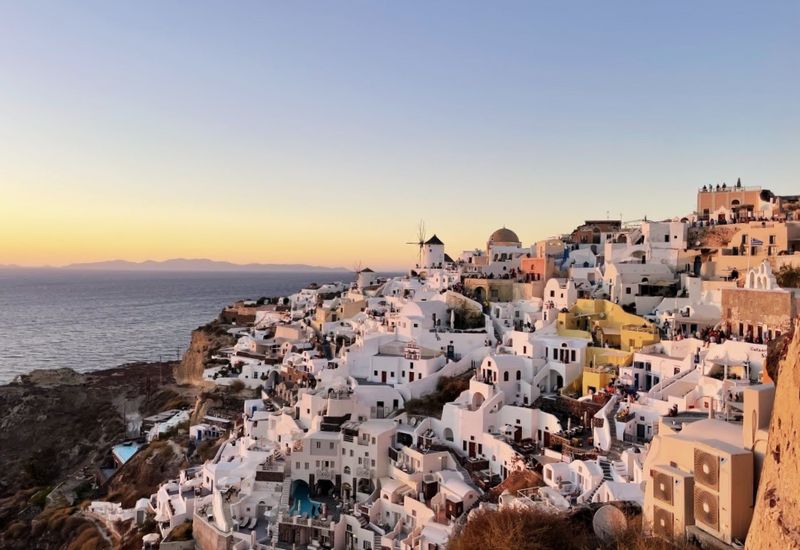 The enchanting evening ambiance of Santorini, Greece, showcasing the iconic whitewashed houses with blue accents perched along the rugged cliffside. As the sun sets, it casts a warm golden hue over the landscape, highlighting the smooth, rounded rooftops and traditional windmills that add to the distinctive aesthetic of the island. The serene Aegean Sea stretches into the horizon, enhancing the tranquil and picturesque setting.