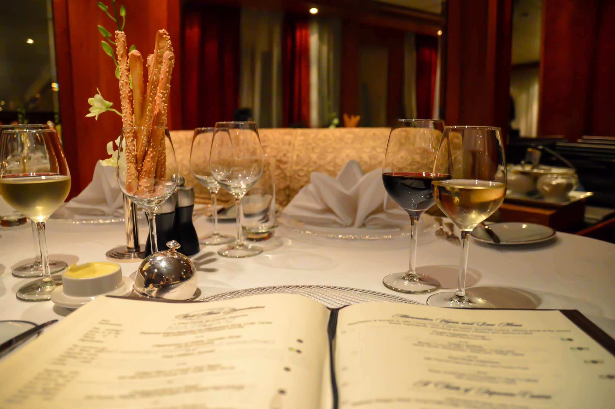 This image depicts an elegantly set dining table, showcasing wine glasses filled with red and white wine as part of a SeaDream wine voyage experience. The table features fine details such as folded white napkins, breadsticks in a tall vase, and a menu in the foreground, with a luxurious interior ambiance in the background. The setting emphasizes refined dining and premium wine pairings.