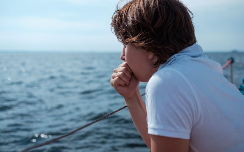 The image captures a young man on a boat, gazing thoughtfully at the ocean. He is wearing a white t-shirt and is holding onto a rope, with the expansive blue sea in the background, suggesting a moment of introspection or seasickness.