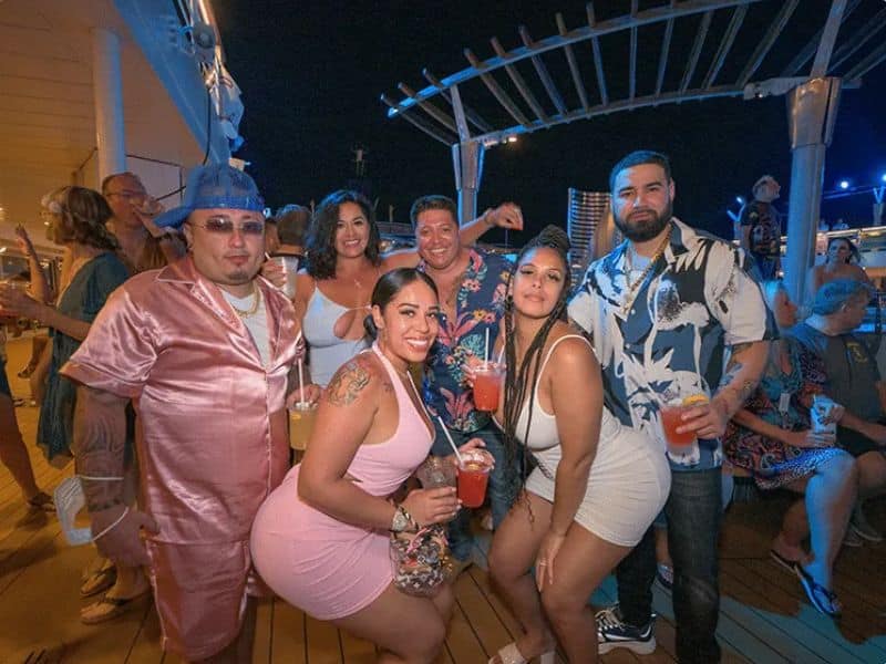 A group of cruise guests enjoying a festive evening on a cruise, with drinks in hand and a lively atmosphere around them.
