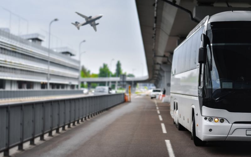 A white shuttle bus is parked at an airport terminal with a plane taking off in the background. The scene is set on a cloudy day, emphasizing the busy travel environment.