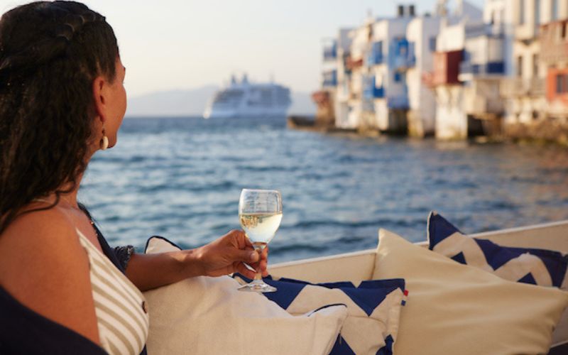 This image captures a Silversea guest enjoying a glass of white wine while overlooking the waterfront in Mykonos. The setting features vibrant blue and white buildings lining the shore, with a cruise ship anchored in the distance under a warm, golden sunset. The scene highlights the serene luxury and picturesque charm of this Greek island destination.