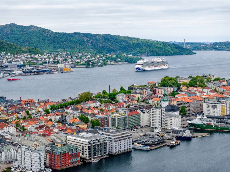 The Sky Princess cruise ship from Princess Cruises sailing near the coast of a vibrant city, with colorful buildings dotting the waterfront and hills in the background.