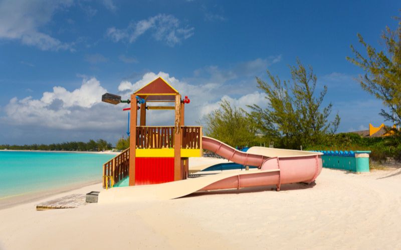 A vibrant children's playground slide sits on a white sandy beach, with the clear turquoise sea providing a picturesque backdrop. The bright colors of the play structure add a sense of fun and leisure to the tranquil beach setting.