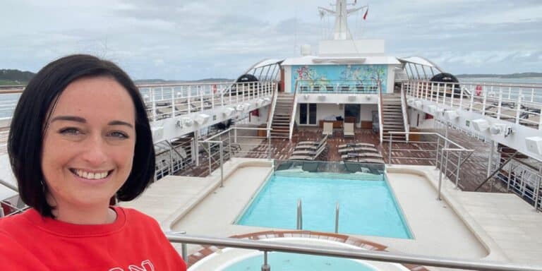 A smiling woman in a red shirt taking a selfie on the top deck of a cruise ship, with a clear view of the pool area and the cloudy sky above.