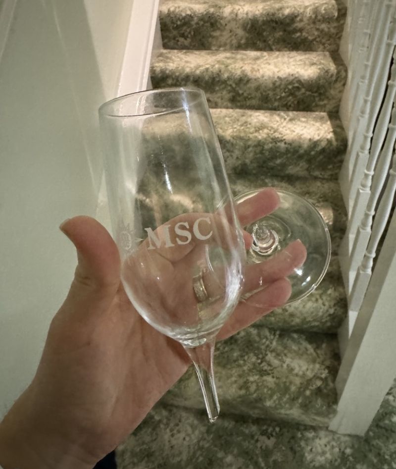 My hand holding an empty MSC Cruises branded champagne glass with a staircase in the background, suggesting the glass is a souvenir kept from a cruise vacation.