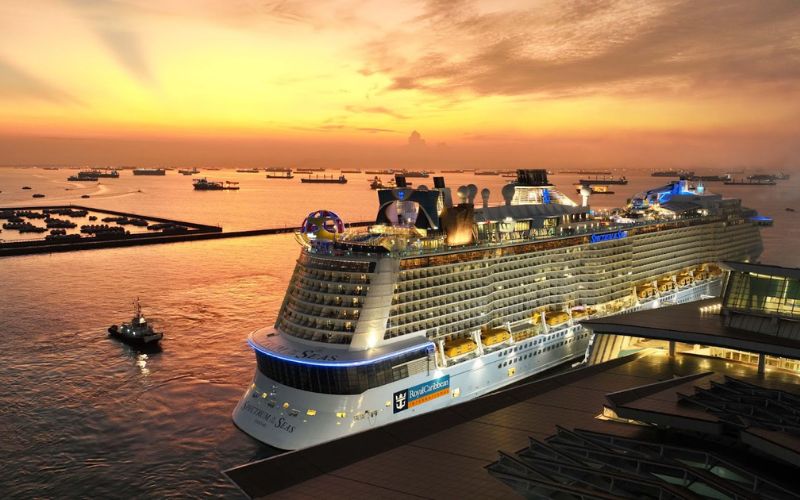 Royal Caribbean's Spectrum of the Seas docked at the port in Singapore during a stunning sunset, with the sky ablaze in warm hues, reflecting on the water filled with ships and boats, emphasizing the ship's impressive size and the port's bustling activity.