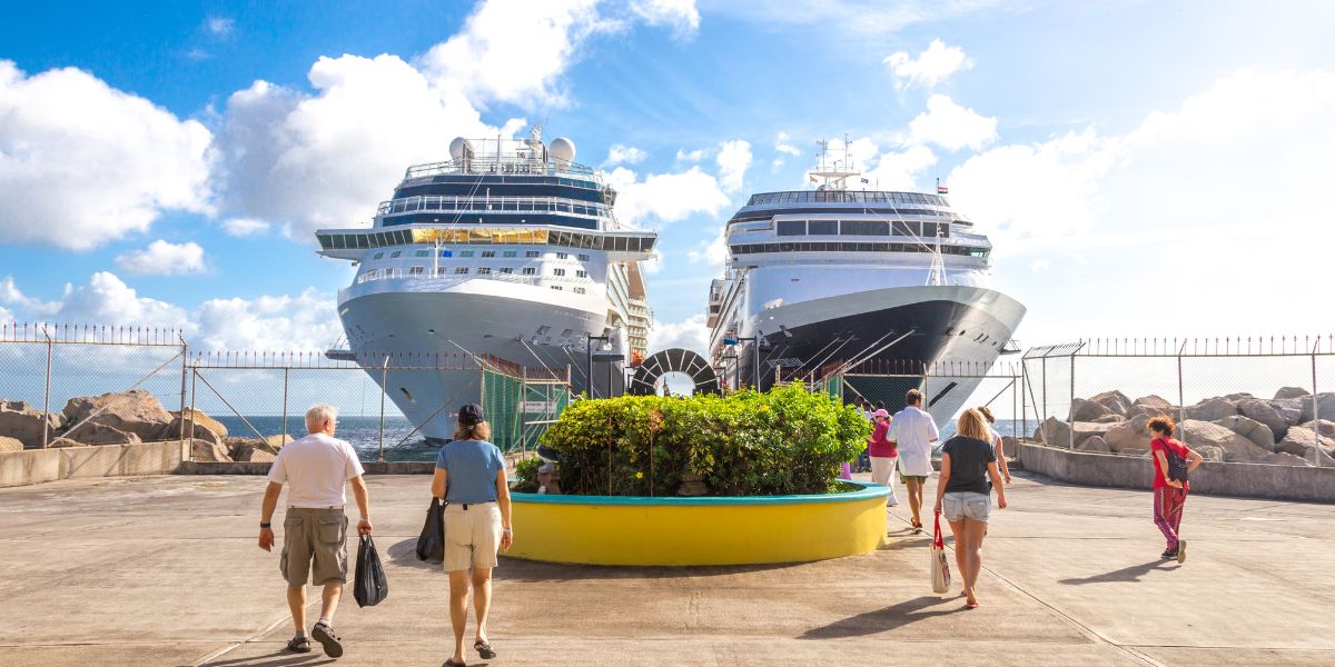 The bustling atmosphere at Port Zante in St. Kitts, with two large cruise ships moored at the terminal. The perspective shows passengers walking along the dock, enjoying the sunny Caribbean weather as they possibly prepare to explore the island or return to the ships after a day out. The ships are impressive in scale, dominating the background, while the foreground features people of various ages in casual vacation attire, emphasizing the tourist-friendly environment. The inclusion of vibrant greenery in a large circular planter adds a touch of the island’s lush natural beauty, contrasting nicely with the industrial elements of the port. This scene effectively portrays Port Zante as a key entry point for visitors to St. Kitts, serving as a gateway to the island's rich culture and scenic landscapes.