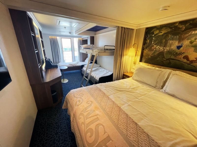 The stateroom on the Disney cruise ship featured in the image is designed to accommodate families comfortably. It includes a large queen-sized bed with elegant bedding and a spacious area for relaxation. Adjacent to the main bed, a wall mural with a whimsical forest theme adds a touch of enchantment, characteristic of Disney's style. The room also features a bunk bed with a top bunk accessible by a ladder, providing additional sleeping space ideal for children. The stateroom is equipped with a desk area and a window to allow for natural light, making the space feel open and welcoming. The overall layout and decor of the room are thoughtfully designed to blend functionality with the magical and fun atmosphere Disney is known for.