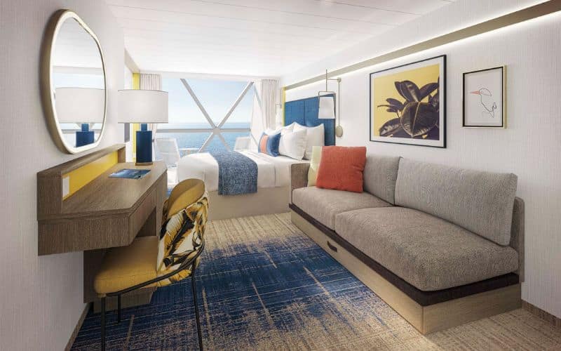 Well-appointed stateroom on Royal Caribbean's 'Star of the Seas' featuring a serene ocean view through a large window. The room is elegantly furnished with a gray sofa, a yellow chair at a small desk, and a comfortable bed adorned with blue and white bedding accented with colorful pillows. Artwork of tropical leaves and minimalist designs add a touch of sophistication to the space.