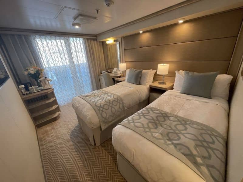 A stateroom on a Princess cruise ship featuring two twin beds with beige and white bedding, a nightstand with a lamp, and a desk. The room has a sliding glass door with patterned curtains leading to a balcony, and a small area with a kettle and flowers on a shelf.