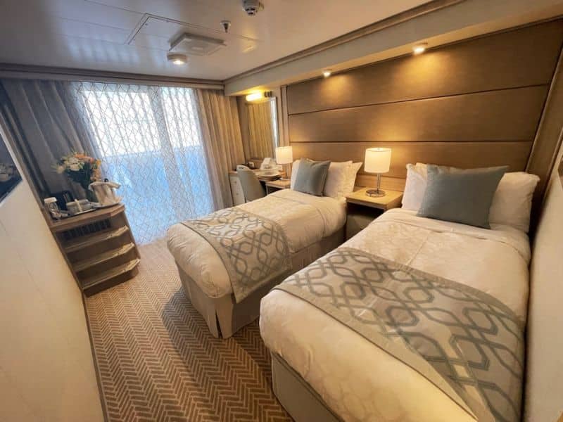 A cozy cruise ship stateroom furnished with two twin beds, patterned throw pillows, and a soft neutral color scheme, with a curtain-drawn window letting in natural light, inviting passengers to a comfortable and restful space at sea.