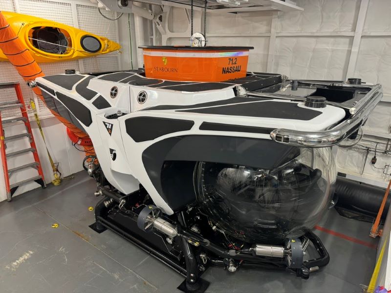 Inside the Seabourn Venture, a sleek white and black submarine is stationed, ready for underwater exploration. This modern submersible features a prominent, clear spherical cabin for viewing, aimed to offer guests an immersive ocean experience. Also visible are safety gear and another submarine in the background, emphasizing the ship's focus on unique guest adventures.