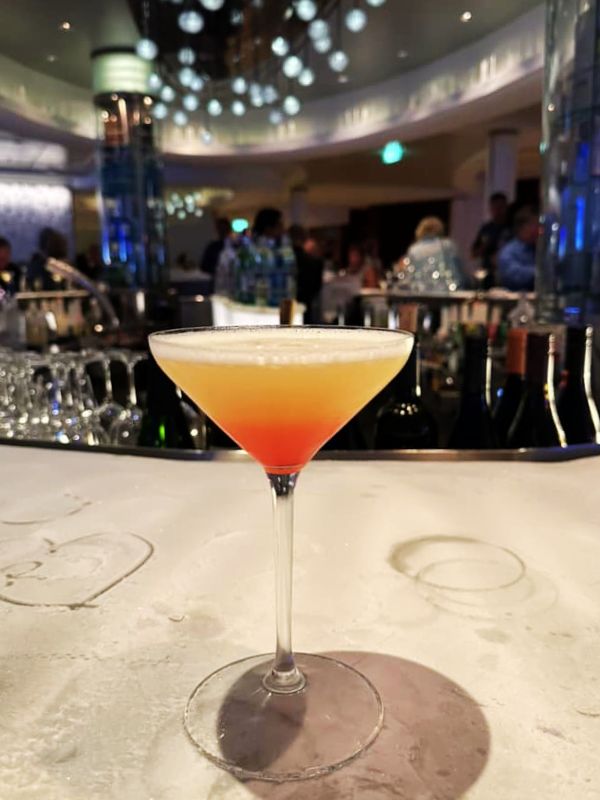A Sunset Martini on a cruise ship, presented in a classic martini glass with a gradient of orange to red, mimicking a sunset. The drink is placed on a bar counter with a sophisticated lounge atmosphere in the background, featuring elegant lighting and patrons enjoying their evening.