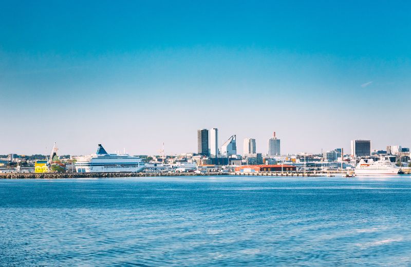 The skyline of Tallinn, Estonia, from the perspective of the sea, featuring a clear view of the city’s diverse architectural styles against a vibrant blue sky. The scene includes modern high-rises, which contrast with the historic and medieval elements that Tallinn is famed for, although these are not visible in this particular shot. A cruise ship is docked at the harbor, indicating the city's popularity as a cruise destination in the Baltic Sea region. The calm waters in the foreground mirror the tranquility of the city, and the overall composition of the photo emphasizes Tallinn's blend of old and new, making it a fascinating city to explore.