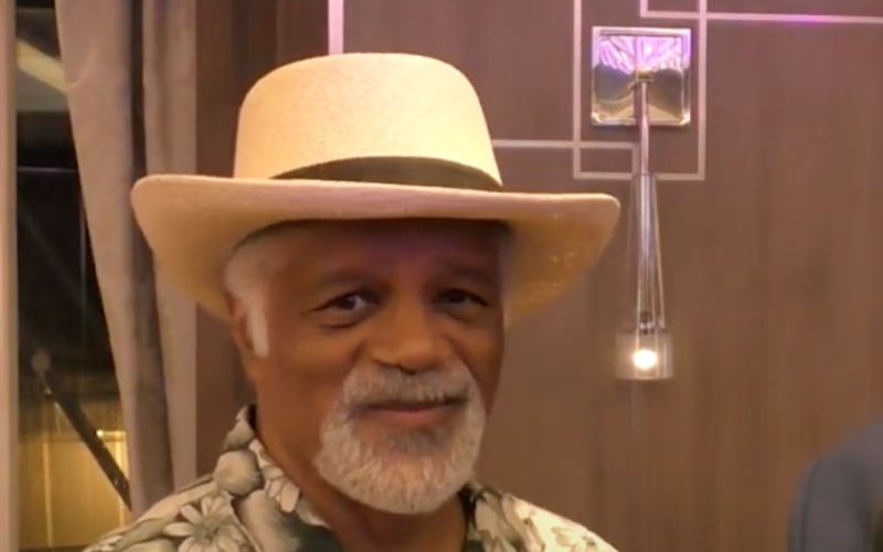 Portrait of Ted Lange with a grey beard, wearing a light-colored fedora and a floral print shirt.
