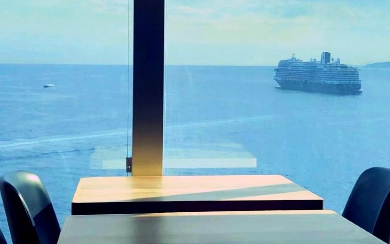 View from a window-side table in a serene room overlooking the sea, with a distant cruise ship sailing along the horizon, capturing a peaceful moment of ocean-view dining or contemplation.