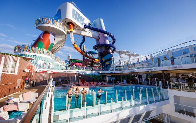 Dynamic view of 'The Hideaway' pool area on Royal Caribbean's 'Icon of the Seas,' bustling with activity. This vibrant deck features a large rectangular pool flanked by pink and white loungers, and is overshadowed by towering, multicolored water slides. Guests enjoy the sun and water in a lively, family-friendly atmosphere.