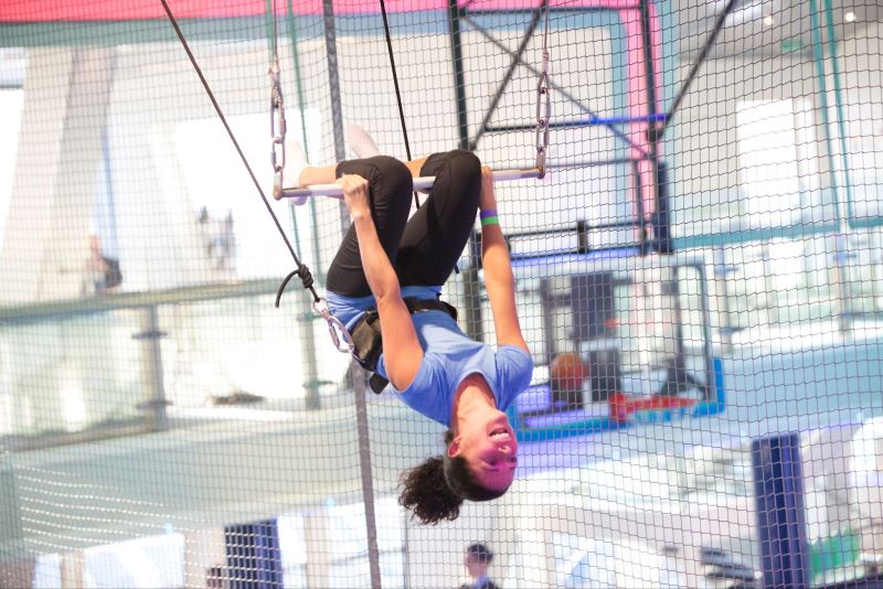 A guest enjoys an exhilarating trapeze experience on Royal Caribbean's Anthem of the Seas, swinging upside down with a secure harness against the indoor activity area's colorful backdrop, showcasing the adventure and entertainment options onboard.