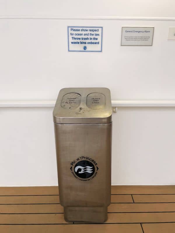 A stainless steel waste bin on a cruise ship labeled for separating 'Paper, Plastic, and Metal' from 'Glass and Cans,' with a sign above it reminding guests to show respect for the ocean and the law by disposing of trash properly onboard.