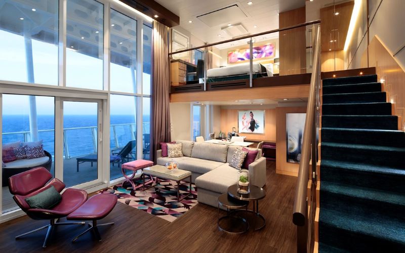 Luxurious two-story cruise ship suite with modern decor, featuring a spacious living area with a large sofa, contemporary furniture, and floor-to-ceiling windows offering an expansive view of the ocean. A sleek staircase leads to the upper-level bedroom, enhancing the room's open and airy feel.