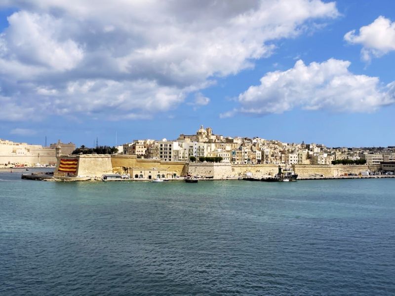 The stunning waterfront of Valletta, the capital city of Malta, viewed from the sea. The historical architecture of Valletta, characterized by sandy-colored stone buildings, is prominently displayed against a backdrop of bright blue skies with fluffy white clouds. The city's dense and layered urban layout rises up from the coastline, featuring various spires and domed rooftops that illustrate its rich history. In the foreground, the clear turquoise waters of the Mediterranean Sea enhance the picturesque setting. A traditional Maltese boat with its distinctive red and yellow stripes adds a touch of local flavor to the scene, highlighting the maritime culture of Malta.