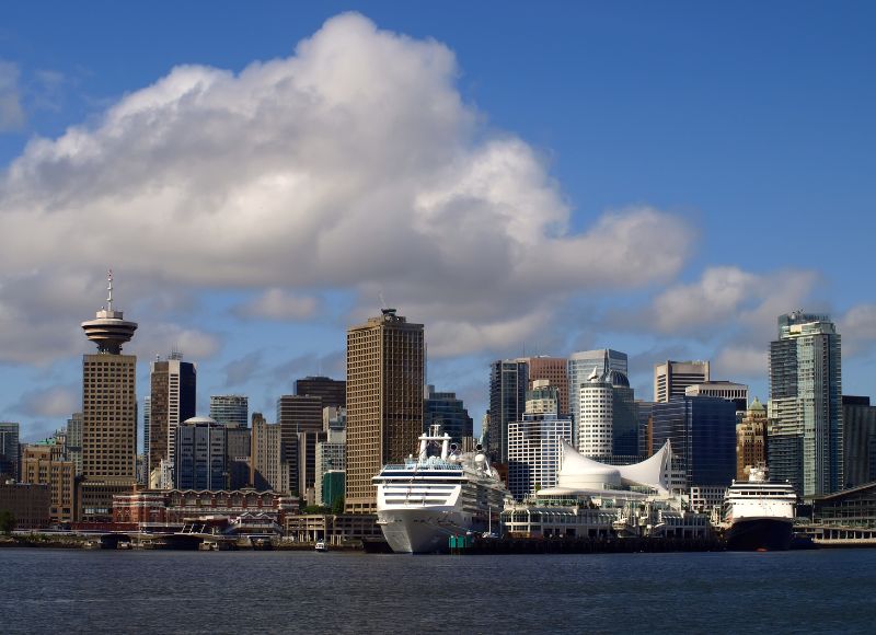 A stunning view of Vancouver, Canada, showcasing its bustling waterfront and distinctive skyline. Prominent in the scene is the iconic Vancouver Lookout tower, offering panoramic views of the city, alongside modern high-rise buildings that define Vancouver's urban landscape. The famous white sails of Canada Place are visible, a landmark that doubles as a cruise ship terminal, underscoring Vancouver's status as a major gateway to the Pacific and a popular stop on numerous cruise itineraries. A cruise ship docked at the terminal adds to the dynamic feel of the harbor, highlighting the city's importance as a maritime hub. The clear skies and fluffy cloud above contribute to the vivid and picturesque setting, capturing the essence of this vibrant coastal city.