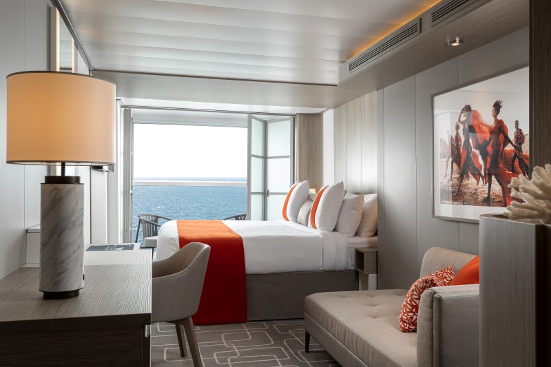 A veranda stateroom on a Celebrity cruise ship elegantly appointed with a comfortable bed accented by an orange throw, a cozy seating area, and a balcony with a view of the ocean, offering guests a private and tranquil retreat.