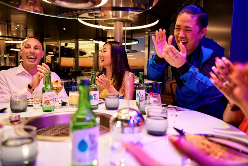 Guests laugh and enjoy an interactive dining experience at the first Korean barbecue restaurant at sea on Virgin Voyages, with soju bottles and grill in the middle of the table, creating a lively and communal mealtime atmosphere.