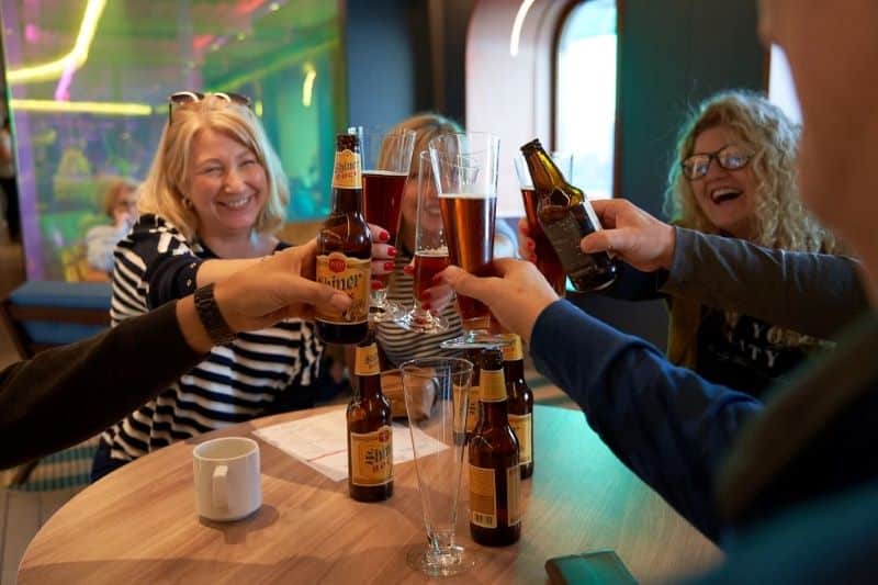 Cheerful guests on Virgin Voyages toast with bottles of Shiner Bock beer, sharing a bucket of beers in a casual and colorful setting, encapsulating the friendly and convivial spirit of the cruise.