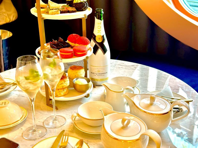An elegant afternoon tea setup on Virgin Voyages with a bottle of Moët & Chandon, delicate pastries, macarons, and finger sandwiches on a tiered server, accompanied by fine china and flutes of champagne, offering a luxurious high tea experience at sea.