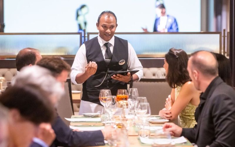 A smiling waiter in a vest and tie taking an order from guests at a well-appointed dining table, with a focus on customer interaction in a sophisticated restaurant setting. A smiling waiter in a vest and tie taking an order from guests at a well-appointed dining table, with a focus on customer interaction in a sophisticated restaurant setting. A smiling waiter in a vest and tie taking an order from guests at a well-appointed dining table, with a focus on customer interaction in a sophisticated restaurant setting.