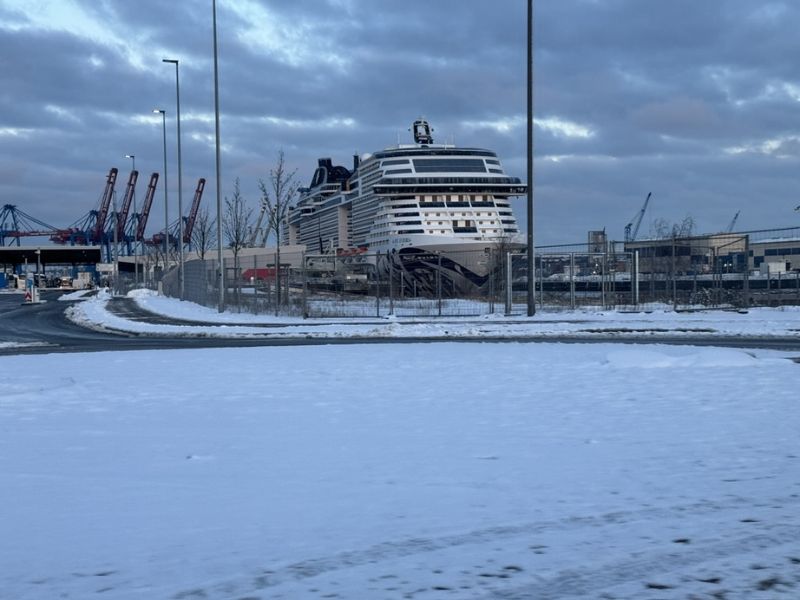 A cruise ship docked at a port during winter, with a snow-covered ground in the foreground and industrial cranes in the background under a cloudy sky. The image conveys a cold, off-season atmosphere possibly contributing to lower cruise prices.