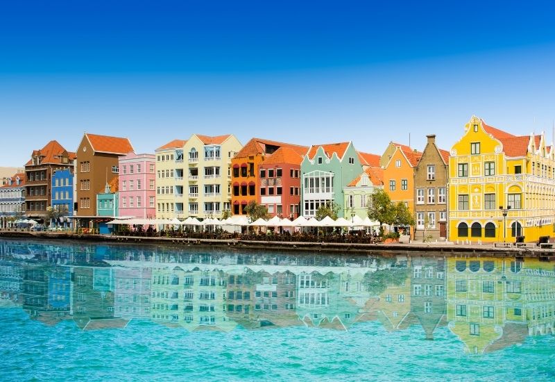 Vibrant and colorful architecture of Willemstad, the capital city of Curaçao. The waterfront is lined with brightly painted buildings that reflect a blend of Dutch colonial architecture and Caribbean flair, making this view instantly recognizable. These historical buildings, with their unique facades and hues ranging from pastel pinks to deep blues and bright yellows, mirror beautifully on the calm waters of the harbor, enhancing the picturesque quality of the scene. Willemstad is not only a UNESCO World Heritage site but also a lively cultural hub, and this image beautifully represents its inviting and cheerful atmosphere.