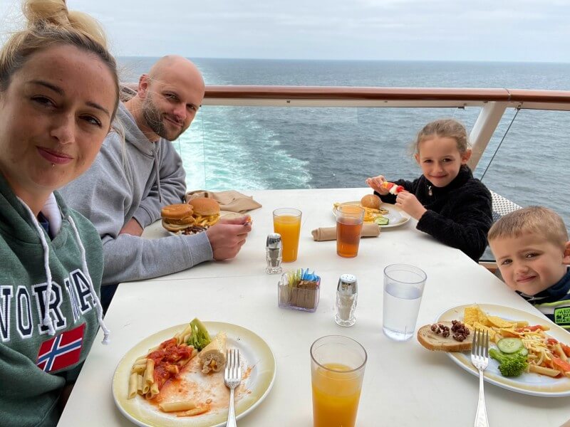 Cruise Mummy and her family are enjoying a meal at an outdoor dining area on a cruise ship, with the ocean in the background. The parents and two children are seated at a table with plates of food, including burgers, fries, pasta, and vegetables, along with glasses of juice and water. They are all smiling and appear to be having a pleasant time together.