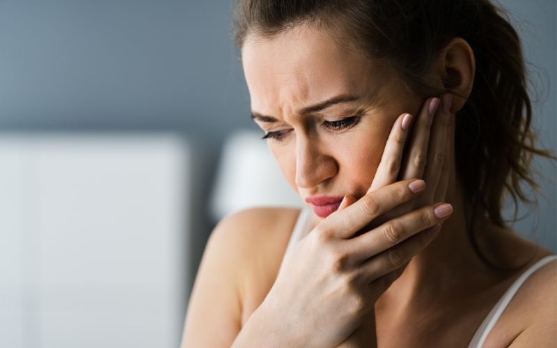  a woman with a troubled expression, holding her cheek in pain, suggesting a toothache or jaw discomfort. She has her hair tied back and is wearing a white tank top, sitting indoors with a blurred background that subtly hints at a home or office environment.