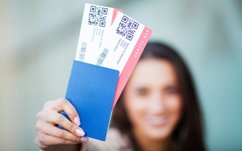 A person holding up a blue passport with two airplane tickets, focusing on the tickets while the person's face is blurred in the background.