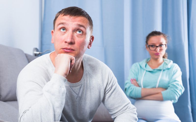 A man in a light gray sweater sits on a couch with a thoughtful expression, resting his chin on his hand, while a woman in a light blue hoodie stands in the background with her arms crossed and a frustrated look on her face. The scene suggests a disagreement or argument between the couple.