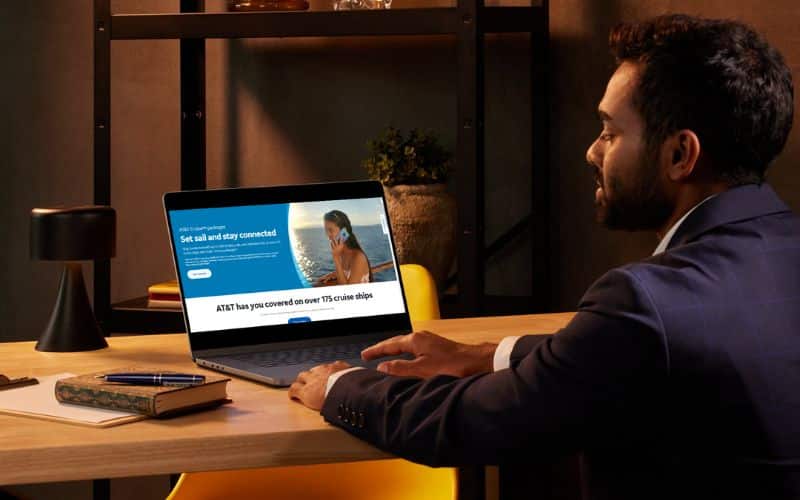 A man in a suit sits at a desk, looking at a laptop screen displaying an AT&T cruise package advertisement. The ad features a woman using a smartphone on a cruise ship deck with the text, "Set sail and stay connected," and "AT&T has you covered on over 175 cruise ships."