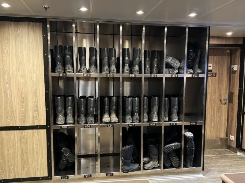 Inside the Seabourn Venture, a neatly organized boot room features rows of black rubber boots, each pair stored in individual compartments labeled with numbers. This room is essential for equipping guests with the necessary footwear for wet landings and rugged terrain during shore excursions. The combination of wood paneling and metal storage units adds a sleek, functional aesthetic to the space.