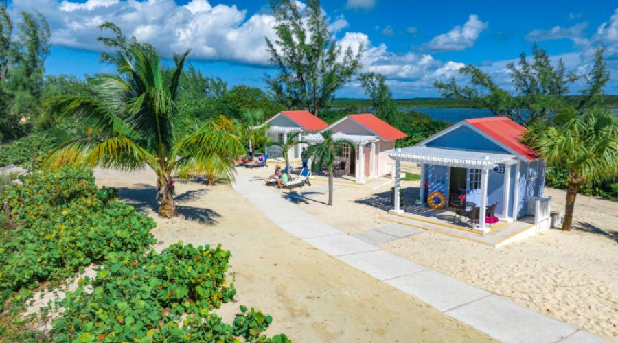 Colorful bungalows line a sandy path, each with a welcoming front porch and vibrant roofing. Palm trees and lush foliage add to the relaxed atmosphere, while guests unwind and savor the island's serene beauty.