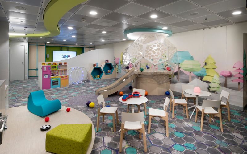 Kids club room on Celebrity Cruise Line, designed with a playful and modern aesthetic. The room features a slide, colorful play structures, small tables and chairs, and a variety of toys, creating an engaging and safe environment for young children to play and explore.