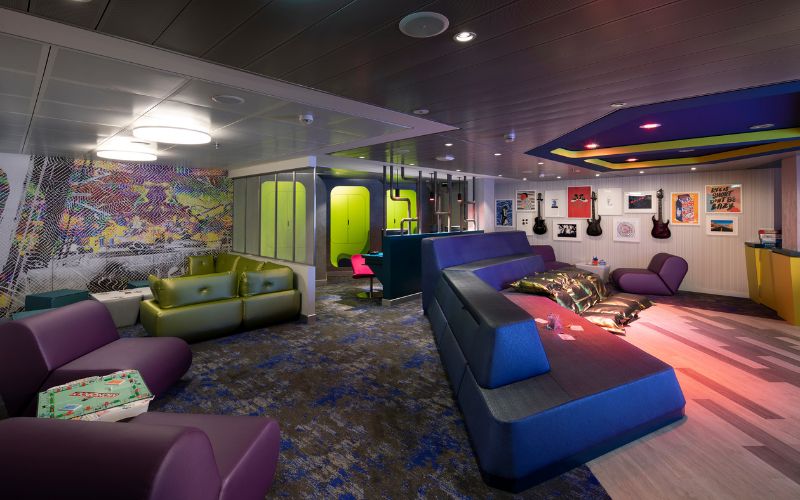 Teens club room on Celebrity Cruise Line, featuring a mix of modern and retro design elements with colorful sofas, wall art, and musical instruments. The room is equipped with various entertainment options and cozy seating areas, providing a trendy and inviting space for teenagers to relax and socialize.