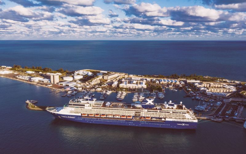 A Celebrity cruise ship docked at a port, captured during the golden hour with a backdrop of scattered clouds in the sky. The waterfront is lined with white buildings and a marina filled with smaller boats, providing a serene coastal setting.