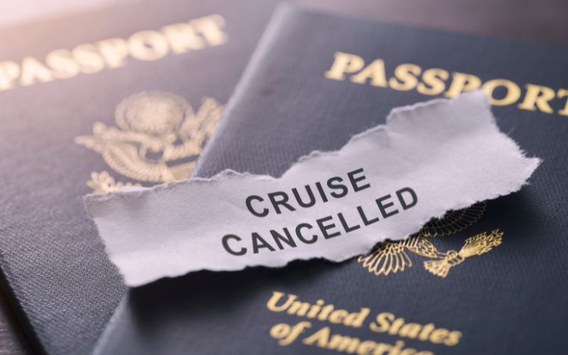 This image depicts two U.S. passports with a torn piece of paper stating "CRUISE CANCELLED" laid across them. The message is a stark, straightforward portrayal of disrupted travel plans, symbolizing disappointment or change in circumstances. The passports and torn paper are shown in close-up with a focus on the text, set against a dark, slightly blurred background to emphasize the central elements.